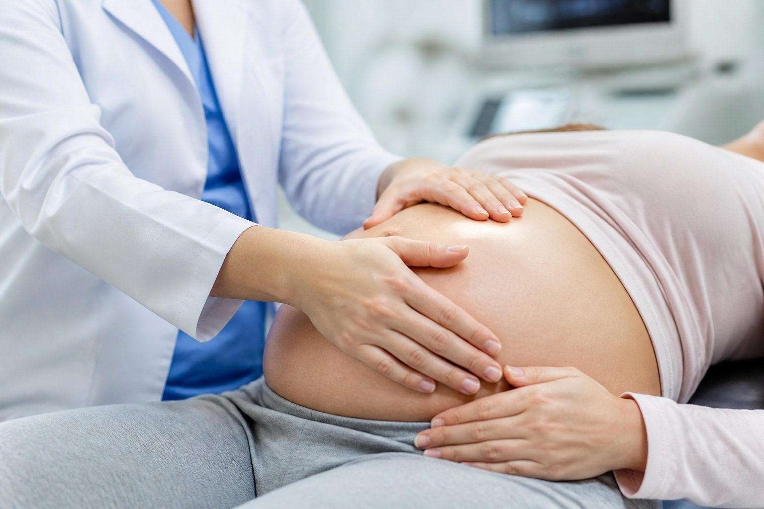 Mãos de um profissional de saúde examinando a barriga de uma mulher grávida em ambiente clínico.